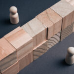 Two wooden peg figures are positioned on opposite sides of a tall wall made of wooden blocks on a dark surface, symbolizing a communication barrier, social divide, or conflict in a business environment