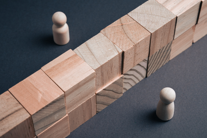 Two wooden peg figures are positioned on opposite sides of a tall wall made of wooden blocks on a dark surface, symbolizing a communication barrier, social divide, or conflict in a business environment